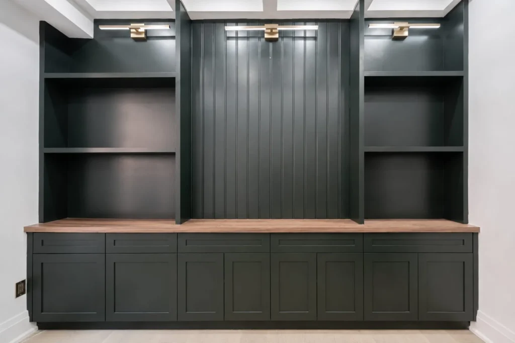 Custom dark green built-in office cabinetry with open shelving, beadboard center panel, brass picture lights and walnut counter